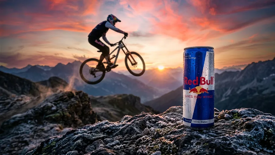 Una lata de Red Bull en primer plano sobre rocas con un ciclista de montaña saltando durante el amanecer al fondo.