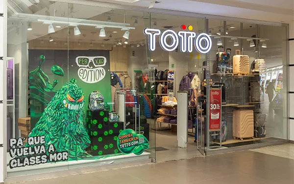 Fachada acristalada de una tienda Totto en un centro comercial, con maletas en exhibición y publicidad escolar.